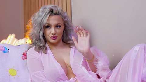Video of a fair-skinned woman with wavy, platinum blonde hair, wearing a sheer, pink, open-front nightgown, sitting on a bed with colorful pillowcases. She waves with a playful expression.