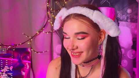 Video of a young woman with long black hair, wearing a white Santa hat, pink eyeshadow, a black choker, and a metal nose ring. Background features festive lights and a wall with graffiti.