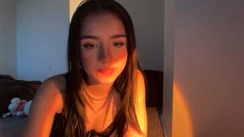 Video of a young woman with long dark hair, wearing a black lace bra, illuminated by a warm light. She has a slender physique, fair skin, and a neutral expression.