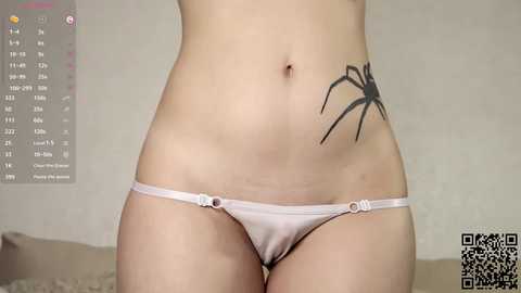 Video of a fair-skinned woman wearing a white thong with a large black spider tattoo on her left hip, partially visible calendar in the background.