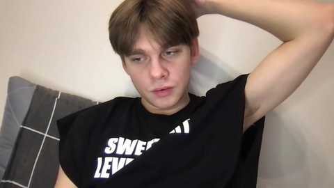 Video of a young, light-skinned man with short brown hair, wearing a black t-shirt with \"SWEAT IS THE EVIL\" text, lying on a grey bed with a white pillow. He has a neutral expression, one arm raised.