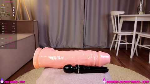 Video of a large, pink, realistic dildo and black vibrator on a white carpet in a modern, minimalist room with grey curtains, wooden floor, and a white dining table.