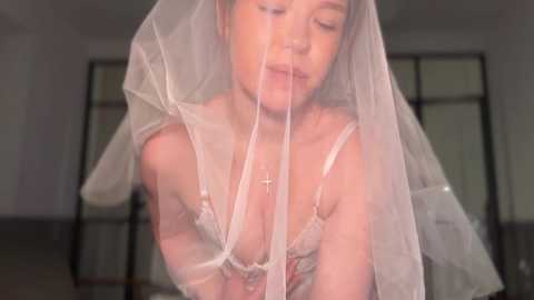 Video of a fair-skinned woman with a slender build wearing a sheer white veil, revealing a white lace bra with a cross necklace. The background is blurred, featuring large windows.