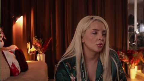 Video of a blonde woman with long hair, wearing a green floral robe, sitting in a dimly-lit room with dark curtains and floral arrangements on a couch.