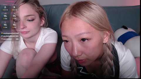Media: Video of two young women with light skin, one with braided blonde hair, one with straight blonde hair, sitting on a couch, looking at a smartphone screen displaying a video feed.
