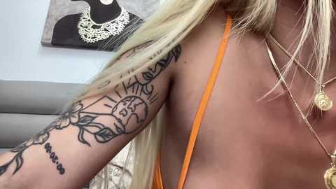 Video of a light-skinned woman with long blonde hair, wearing an orange halter top, showcasing multiple tattoos on her left arm, including a sunflower and a face. She has a gold necklace and is indoors with a gray couch in the background.