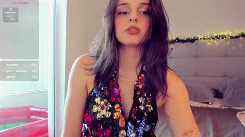 Media: Video of a young woman with long dark hair, wearing a black halterneck dress adorned with colorful sequins, standing indoors near a bed with a gray tufted headboard.