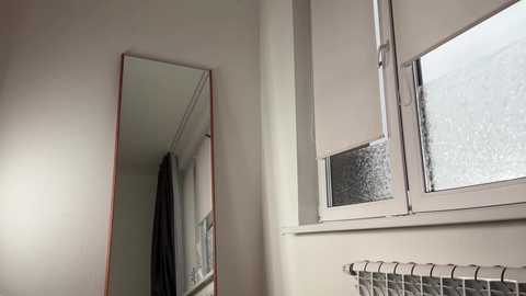 A video of a minimalist, modern bathroom with a large, rectangular mirror mounted on a white wall. The mirror reflects a partially visible shower curtain and a frosted window with white blinds. The room is well-lit, with soft, natural light filtering through the window.