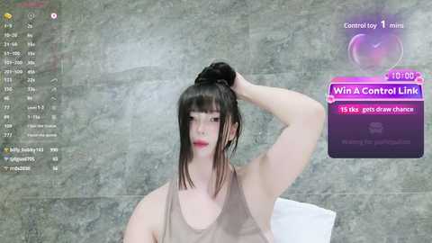 Video of a young Asian woman with fair skin, wearing a sheer beige tank top, with straight black hair styled in a bun. Background features a gray marble wall, and a screen display reads \"Win a Control Link\" with a 10:02 time.