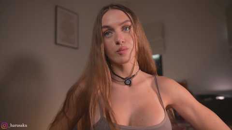 Media: Video of a Caucasian woman with long, straight, light brown hair, wearing a beige tank top, standing indoors. She has a slender physique and wears a black choker necklace. Background features a blurred, dimly lit room with framed artwork.