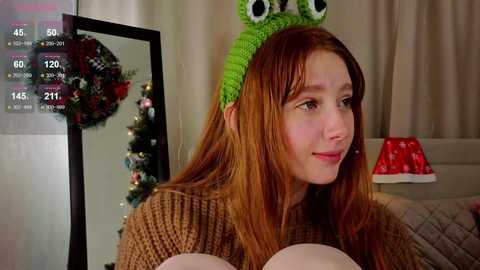 A video of a young woman with long red hair, wearing a green frog headband and a brown sweater, sitting indoors, looking at a digital display. Christmas decorations are in the background.