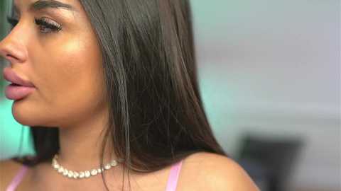 Video of a woman with long, straight, dark brown hair and light skin, wearing a pink spaghetti-strap top and a pearl necklace. Background is blurred, featuring a hint of green and white walls.