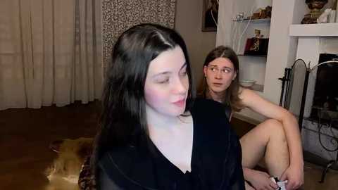 Video of two young women in a cozy living room: one with pale skin and long black hair, wearing a black dress, sitting on a couch; the other with fair skin, brown hair, and a black top, sitting on the floor.