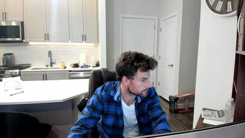 A video of a man with short dark hair in a blue plaid shirt, leaning against a glass wall in a modern kitchen with white cabinets, stainless steel appliances, and a clock on the wall.