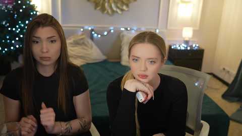 Media: Video of two young women with fair skin, one with long brown hair and tattoos, the other with blonde hair, seated at a table, looking contemplative in a cozy, decorated room with a lit Christmas tree.