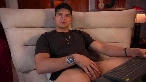 Video of a young Asian man with short black hair, wearing a dark t-shirt and no pants, sitting on a beige sofa, holding a remote, in a dimly lit living room.