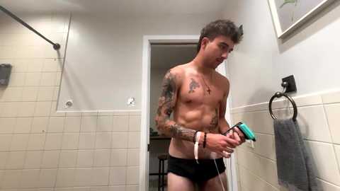 Video of a muscular, shirtless man with tattoos, wearing black briefs, using a hair dryer in a beige-tiled bathroom.