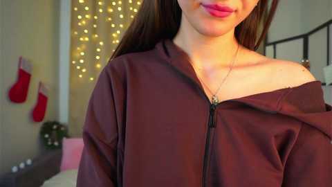 Video of a smiling woman with long brown hair, wearing a dark brown off-shoulder top with a zipper, standing in a warmly lit, festive room with Christmas decorations.