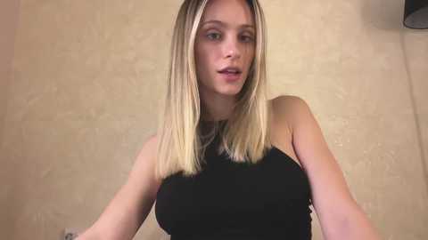 Video of a young, fair-skinned woman with long, straight blonde hair, wearing a black sleeveless top, against a beige, textured wall background.