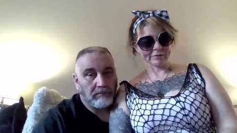 A video of a bald, bearded man and a blonde woman with glasses and a headband, both wearing fishnet tops, indoors.