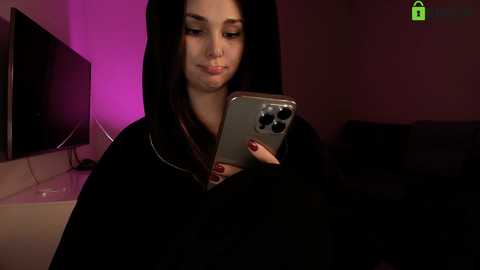 Video of a young woman with fair skin, long black hair, wearing a black robe, holding a silver iPhone in a dimly lit room with a purple accent light and a TV in the background.