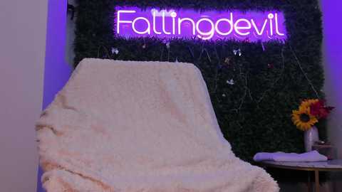 Video of a cozy, dimly lit room with a plush white blanket draped over a chair, a vibrant sunflower on a table, and a neon sign reading \"Fattynoodle\" against a lush green wall.