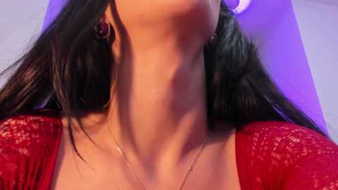Video of a woman with fair skin, long black hair, wearing a red lace top, lying back, head tilted up, purple lighting, neck visible.