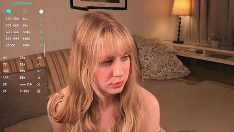 Media: A video of a blonde woman with long hair, seated on a beige sofa in a living room, wearing a strapless dress, with a digital display overlay showing heart rate and other health metrics.