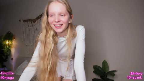 A young, fair-skinned girl with long, wavy blonde hair, wearing a white long-sleeve shirt, smiles brightly in a cozy, dimly lit room with a macrame wall hanging, green plant, and \"25 love\" text overlay.