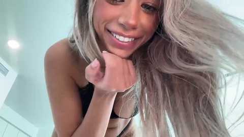 Media: A close-up video of a smiling woman with long, wavy blonde hair, wearing a black strapless top. She's playfully pulling her hair with her left hand. The background is a bright, modern room with white walls and ceiling.