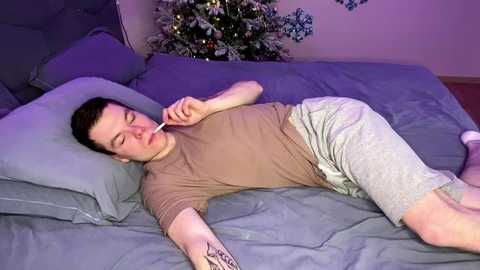 Video of a pale, young man with short brown hair, lying on a bed in a light gray shirt and shorts, with a thermometer in his mouth, surrounded by festive decor.