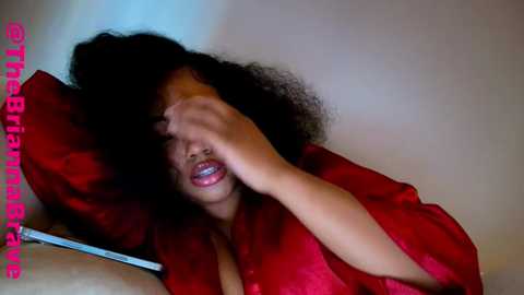 Video of a young woman with dark skin and curly hair, partially covered with a red satin robe, lying on a beige couch, looking distressed.