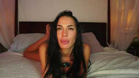 Video of a topless woman with long black hair lying on a bed, making a pouty face, in a dimly lit room with sheer curtains and a white lamp.