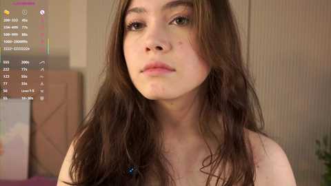 Media: Video of a young Caucasian woman with long, wavy brown hair, fair skin, and a slight pout. She has a few acne spots on her cheeks. The background is a beige room with a computer screen displaying text and icons.