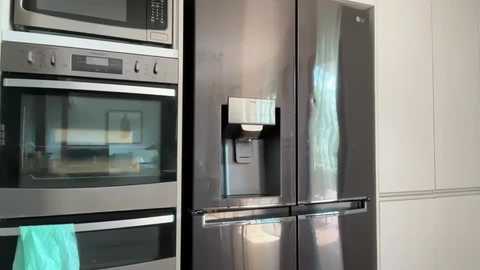 A video of a modern kitchen with stainless steel appliances: a double-door refrigerator, built-in microwave, and oven, all sleek and shiny, against white cabinets. A turquoise hand towel hangs on the oven door handle.