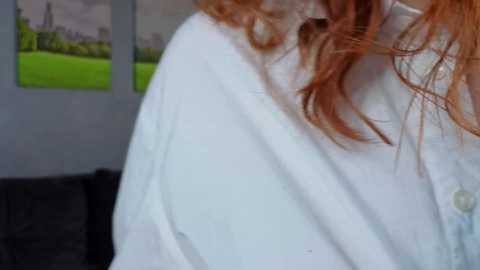 Video of a woman with long, wavy red hair wearing a white robe, standing in a room with green grass artwork on the wall and a dark couch in the background.