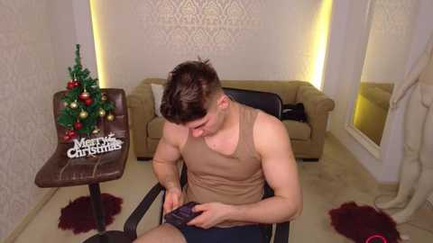 Video of a muscular, shirtless man in beige shorts, seated on a chair, adjusting his phone, in a cozy, dimly-lit living room with a Christmas tree, a beige sofa, and decorative rugs.