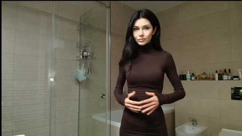 Video of a slender, dark-haired woman in a tight, long-sleeved brown dress, holding her hands together, standing in a modern, beige-tiled bathroom with a glass shower and various toiletries on shelves.