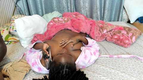 Video of a nude Black woman with braids, lying on a bed covered in a pink and white patterned blanket, holding a phone to her ear. Surrounding her are various colorful pillows and a gray curtain background.