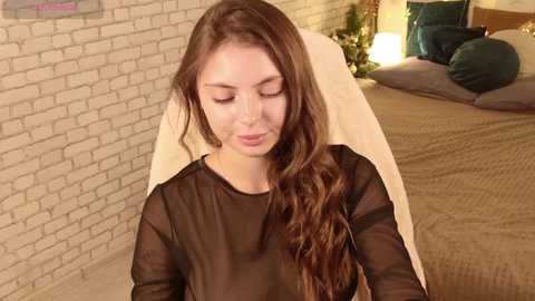 Video of a young woman with long brown hair, wearing a sheer black top, sitting on a beige chair in a cozy bedroom with a brick wall, pillows, and a green lamp.