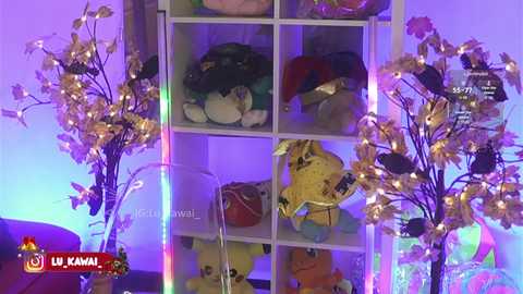 Video of a toy-filled white bookshelf in a room with purple and blue lighting. Toys include plush animals, a yellow horse, and a red car. Artificial floral branches with fairy lights adorn the sides.