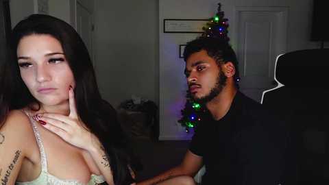 Video of a distraught woman with long dark hair and a tattooed arm, sitting next to a calm man with short dark hair, in a dimly lit living room with a decorated Christmas tree.