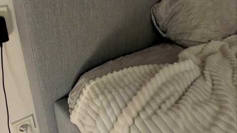 A video of a corner of a gray fabric sofa with a white, ribbed blanket draped over it, revealing a textured, patterned cushion underneath. An electrical outlet is visible on the wall in the background.