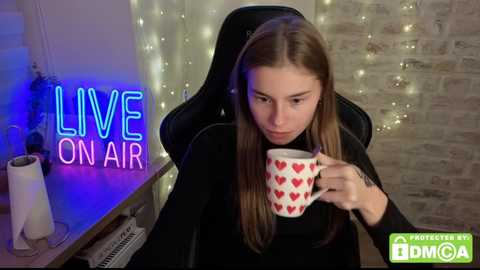 A video of a young woman with long brown hair in a black top, sipping from a red heart-patterned mug. Text reads \"LIVE ON AIR\" in neon blue and pink.