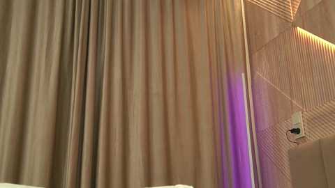 Video of beige, pleated curtains partially drawn, revealing a modern, geometrically patterned wall with warm, ambient lighting. A small white device is mounted on the wall near the curtain.