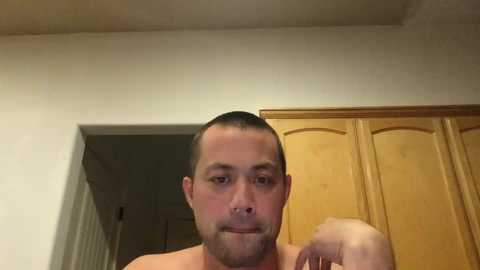 A video of a shirtless man with short brown hair and a trimmed beard, wearing a nose ring, standing indoors in front of a wooden closet door.