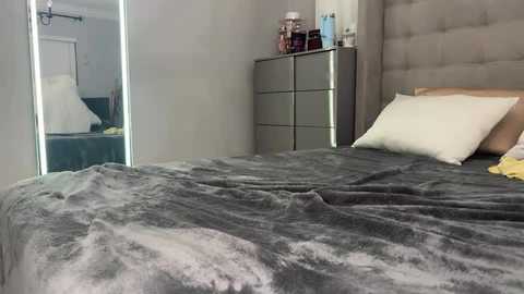 Video of a modern bedroom with a gray, textured comforter, white pillows, and a plush gray upholstered headboard. A mirrored closet door reflects the room, and a dresser with toiletries and a lamp stands against the wall.