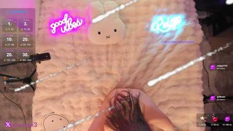 A video captures a person with long hair, lying on a plush bed, with neon lights and a \"good vibes\" sign in the background.