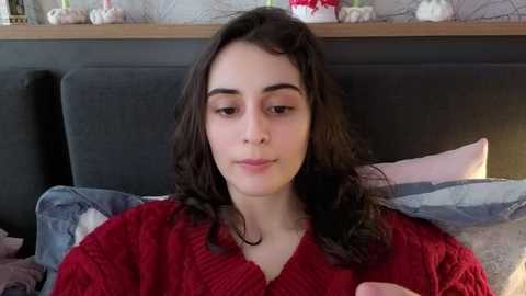 Media: Video of a young woman with long, curly brown hair, fair skin, and a red knit sweater, sitting on a bed with patterned pillows.