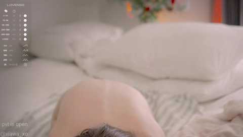Video of a naked person lying face down on a bed with white sheets and pillows, blurred background showing a green plant and a window.